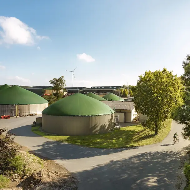 Biogasanlage mit mehreren grünen Silos, umgeben von Bäumen und blühender Natur, unter einem sonnigen Himmel. Eine Person fährt mit dem Fahrrad auf der Straße, die das nachhaltige und umweltfreundliche Konzept unterstreicht.