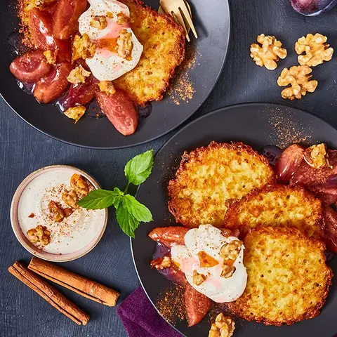 Zwei elegant angerichtete Teller mit knusprigen Mini-Reibekuchen, serviert mit karamellisiertem Zwetschgenröster, cremigem Joghurt und gerösteten Walnüssen. Die goldbraunen Kartoffelpuffer liegen auf dunklen Tellern, begleitet von goldfarbenem Besteck auf einer violetten Stoffserviette. Frische Zwetschgen, Zimtstangen und Walnüsse als Dekoration betonen die herbstliche Atmosphäre des Gerichts.