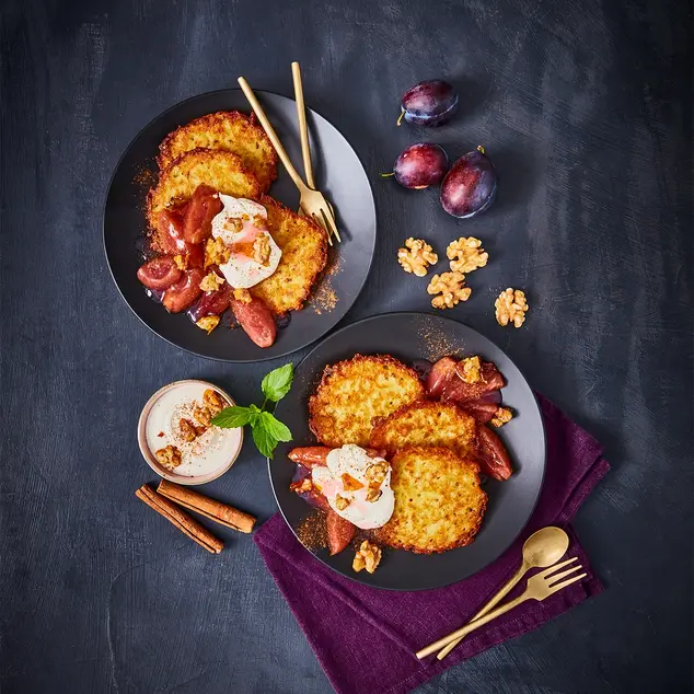 Zwei elegant angerichtete Teller mit knusprigen Mini-Reibekuchen, serviert mit karamellisiertem Zwetschgenröster, cremigem Joghurt und gerösteten Walnüssen. Die goldbraunen Kartoffelpuffer liegen auf dunklen Tellern, begleitet von goldfarbenem Besteck auf einer violetten Stoffserviette. Frische Zwetschgen, Zimtstangen und Walnüsse als Dekoration betonen die herbstliche Atmosphäre des Gerichts.