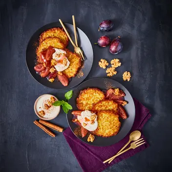 Zwei elegant angerichtete Teller mit knusprigen Mini-Reibekuchen, serviert mit karamellisiertem Zwetschgenröster, cremigem Joghurt und gerösteten Walnüssen. Die goldbraunen Kartoffelpuffer liegen auf dunklen Tellern, begleitet von goldfarbenem Besteck auf einer violetten Stoffserviette. Frische Zwetschgen, Zimtstangen und Walnüsse als Dekoration betonen die herbstliche Atmosphäre des Gerichts.