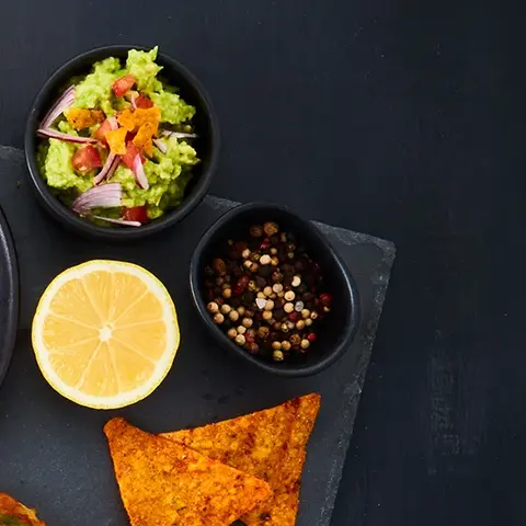 Eine stilvolle Präsentation von Mini-Reibekuchen auf einer dunklen Schieferplatte. Die Reibekuchen sind abwechslungsreich garniert, unter anderem mit Guacamole, Lachs, Kräuterschmand und Camembert mit Preiselbeeren. Begleitend sind kleine Schälchen mit Toppings wie Guacamole, Pfefferkörnern, Preiselbeeren und Zitrone sowie Tortilla-Chips arrangiert. 