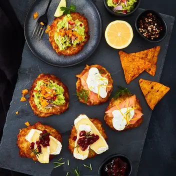 Eine stilvolle Präsentation von Mini-Reibekuchen auf einer dunklen Schieferplatte. Die Reibekuchen sind abwechslungsreich garniert, unter anderem mit Guacamole, Lachs, Kräuterschmand und Camembert mit Preiselbeeren. Begleitend sind kleine Schälchen mit Toppings wie Guacamole, Pfefferkörnern, Preiselbeeren und Zitrone sowie Tortilla-Chips arrangiert. 