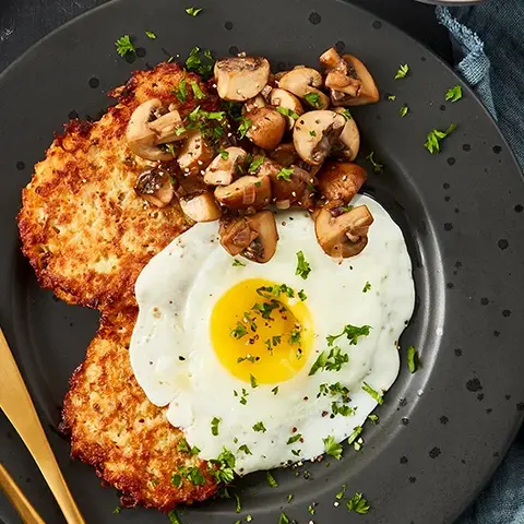 Schinken-Rösti angerichtet mit einem Spiegelei und sautierten Champignons, serviert auf einem dunklen Teller. Beilage: frischer grüner Blattsalat in einer Schale, garniert mit Petersilie und Salzflocken. Dekoriert mit goldfarbenem Besteck und einem blauen Leinentuch.