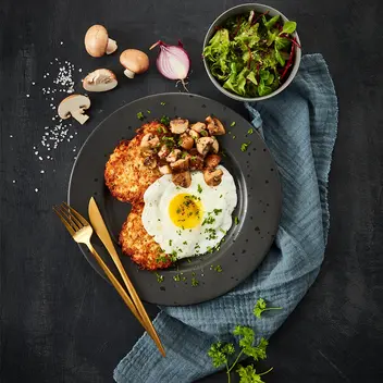 Schinken-Rösti angerichtet mit einem Spiegelei und sautierten Champignons, serviert auf einem dunklen Teller. Beilage: frischer grüner Blattsalat in einer Schale, garniert mit Petersilie und Salzflocken. Dekoriert mit goldfarbenem Besteck und einem blauen Leinentuch.