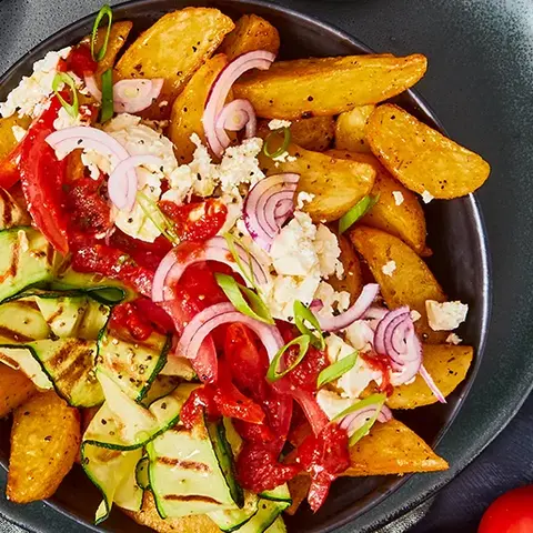 Krosse Wedges-Bowl mit frischem Zucchinisalat, saftigen Tomaten, roten Zwiebeln und einer Prise Feta, serviert mit einem würzigen Dip. Angerichtet auf einem dunkelgrauen Teller mit grüner Stoffserviette, begleitet von frischen Tomatenrispen.