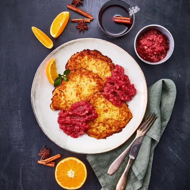 Drei knusprige Kartoffel-Reibekuchen auf einem weißen Teller, serviert mit einem fruchtigen Glühwein-Apfelmus. Dekoriert mit einer frischen Orangenscheibe und Minzblättern. Im Hintergrund Gewürze wie Zimtstangen, Sternanis und Orangenscheiben sowie eine Tasse dampfender Glühwein mit einer Zimtstange. Das Besteck liegt auf einer olivgrünen Stoffserviette, arrangiert auf einem dunklen Untergrund.