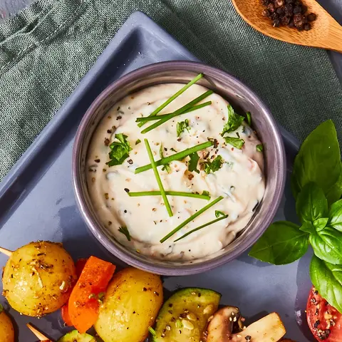 Gegrillte Rosmarinkartoffel-Gemüse-Spieße auf einer grauen Servierplatte, belegt mit Kartoffeln, Zucchini, Paprika, Champignons und Tomaten, garniert mit frischen Kräutern. Daneben eine Schale mit Kräuterquark, dekoriert mit Schnittlauch. Das Arrangement ist auf einer grauen Holzunterlage mit einer grünen Stoffserviette präsentiert. Im Hintergrund liegen frischer Basilikum, Pfefferkörner und ein Holzlöffel, alles auf einem dunklen Hintergrund.