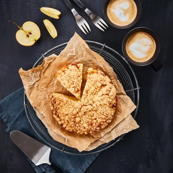 Ein frisch gebackener Apfelkuchen mit Streuseln, serviert auf Backpapier und einem Gitter. Daneben liegen Apfelscheiben, zwei Kaffeetassen mit Latte Art und Gabeln auf einem dunklen Hintergrund.