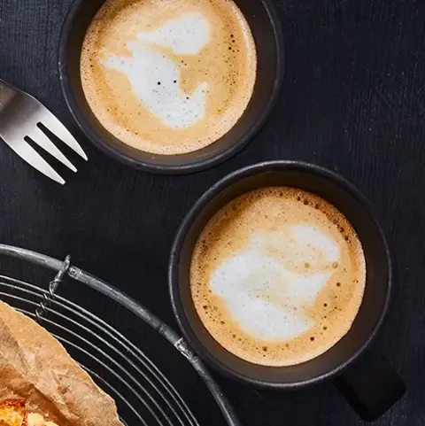 Ein frisch gebackener Apfelkuchen mit Streuseln, serviert auf Backpapier und einem Gitter. Daneben liegen Apfelscheiben, zwei Kaffeetassen mit Latte Art und Gabeln auf einem dunklen Hintergrund.