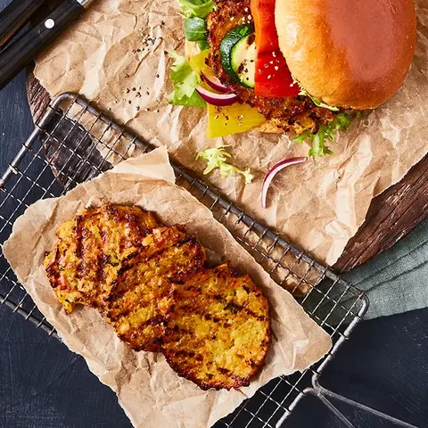 Ein vegetarischer Burger mit Gemüsegrillis als Patty, frischem Salat, Gurken, Tomaten und Zwiebeln, serviert auf Backpapier und einem rustikalen Holzbrett. Daneben liegt ein Gitter mit weiteren gegrillten Gemüsegrillis sowie ein kleines Schälchen mit Dip. Besteck befindet sich ebenfalls auf dem Holzbrett, alles auf einem dunklen Hintergrund.