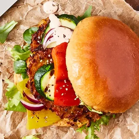 Ein vegetarischer Burger mit Gemüsegrillis als Patty, frischem Salat, Gurken, Tomaten und Zwiebeln, serviert auf Backpapier und einem rustikalen Holzbrett. Daneben liegt ein Gitter mit weiteren gegrillten Gemüsegrillis sowie ein kleines Schälchen mit Dip. Besteck befindet sich ebenfalls auf dem Holzbrett, alles auf einem dunklen Hintergrund.