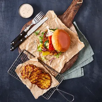 Ein vegetarischer Burger mit Gemüsegrillis als Patty, frischem Salat, Gurken, Tomaten und Zwiebeln, serviert auf Backpapier und einem rustikalen Holzbrett. Daneben liegt ein Gitter mit weiteren gegrillten Gemüsegrillis sowie ein kleines Schälchen mit Dip. Besteck befindet sich ebenfalls auf dem Holzbrett, alles auf einem dunklen Hintergrund.