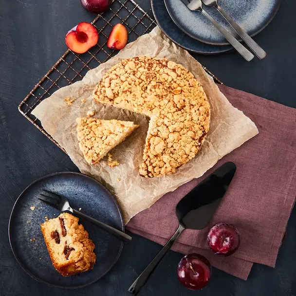 Ein Pflaumenkuchen mit Streuseln, serviert auf Backpapier und einem Gitter, ein Stück wurde herausgeschnitten. Auf einem kleinen Teller liegt ein Stück des Kuchens mit sichtbaren Pflaumenstücken, daneben eine Gabel. Frische, halbierte und ganze Pflaumen dienen als Dekoration. Im Hintergrund stehen zwei leere Teller mit Gabeln, alles auf einem dunklen Hintergrund mit einer violetten Stoffserviette arrangiert.