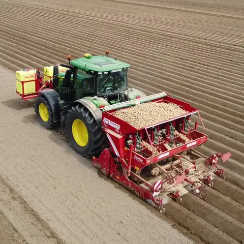 Ein moderner Traktor zieht eine Kartoffelsetzmaschine über ein weites, frisch gepflügtes Feld. Die Maschine ist mit Kartoffeln gefüllt und setzt diese in die vorbereiteten Furchen des Feldes. Die Reihen verlaufen parallel und erstrecken sich über die gesamte Fläche.