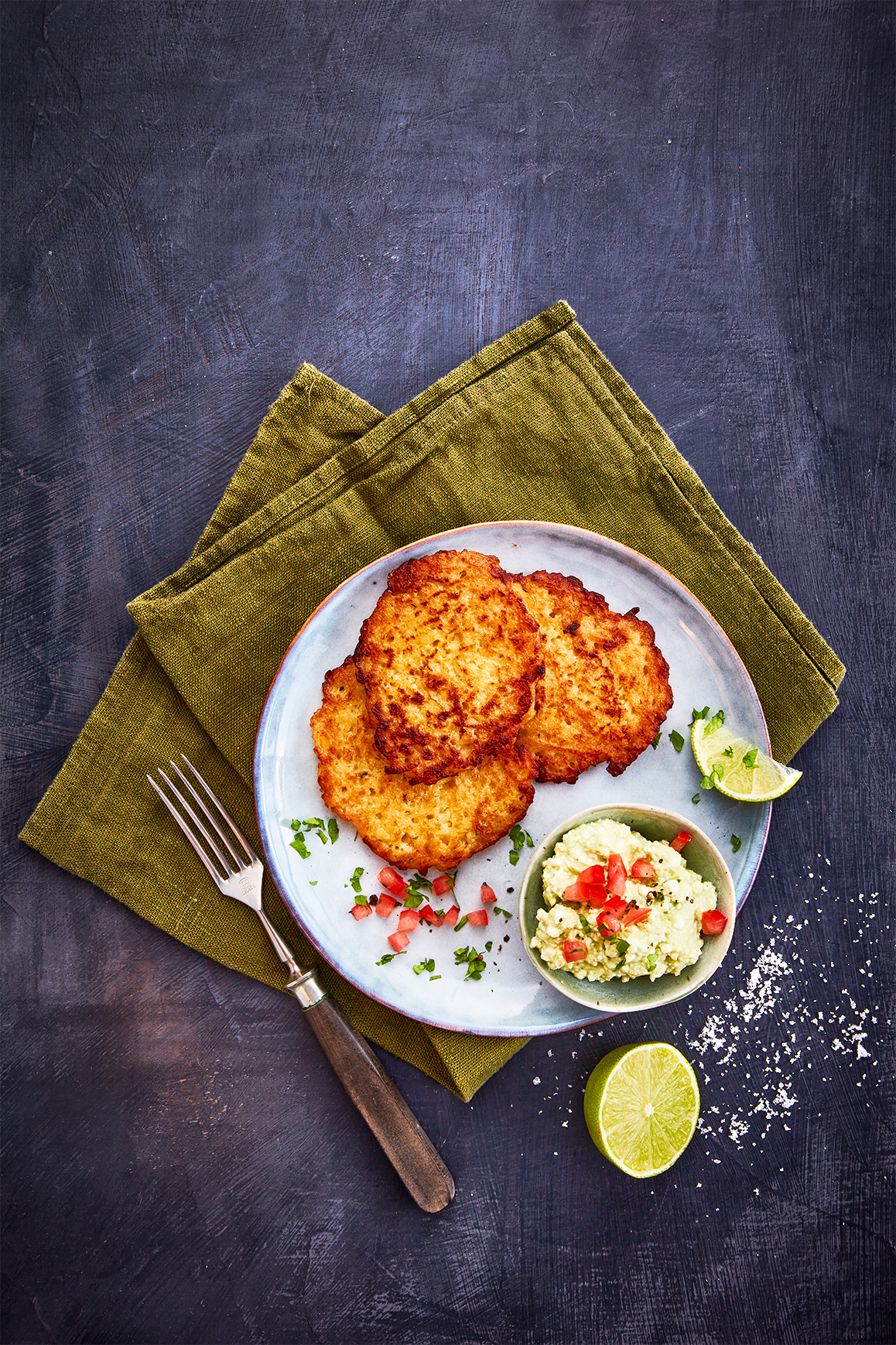 Ein Teller mit drei knusprigen Reibekuchen, serviert mit einer kleinen Schale Avocadocreme, garniert mit gehackten Tomaten und frischen Kräutern. Eine halbe Limette und grobes Meersalz liegen dekorativ daneben. Das Arrangement ist auf einer olivgrünen Stoffserviette präsentiert, begleitet von einer Gabel mit Holzgriff, alles auf einem dunklen Hintergrund.