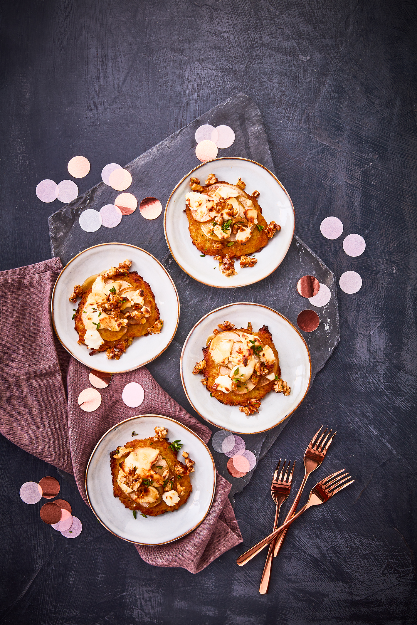 Vier Teller mit Mini-Reibekuchen, belegt mit Birnenscheiben, geschmolzenem Ziegenkäse und karamellisierten Walnüssen, garniert mit frischen Kräutern. Das Arrangement ist auf einer dunklen Schieferplatte angerichtet, dekoriert mit roséfarbenen Konfetti und einer violetten Stoffserviette. Daneben liegen roséfarbene Gabeln, alles präsentiert auf einem dunklen Hintergrund.